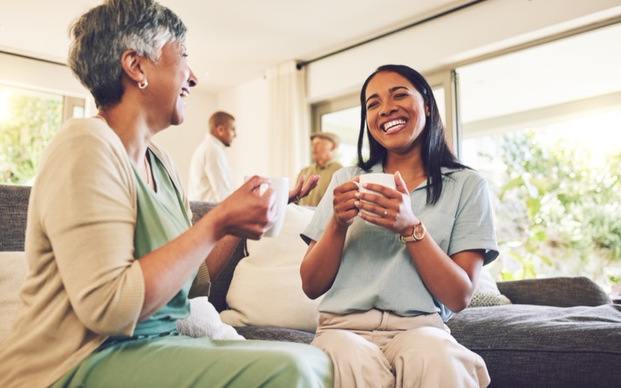 An older woman and her caregiver laughing together over a cup of tea