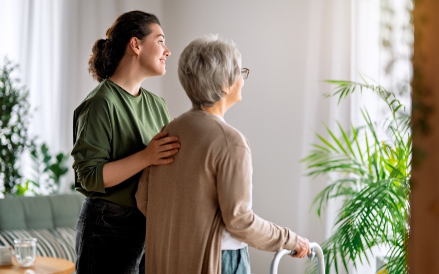 A caregiver supporting an older woman as she walks with a wheeled walker