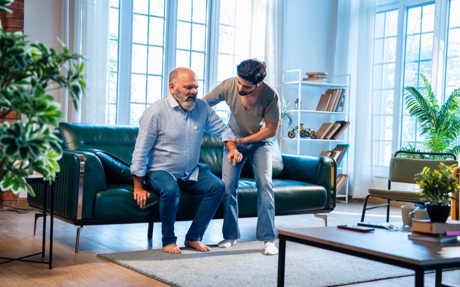 A caregiver steadying an older man as he stands up from the sofa at home