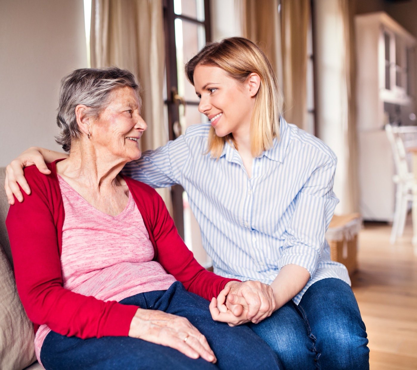 A Home Haven caregiver sitting on the sofa beside an older client, holding her hand and smiling with her