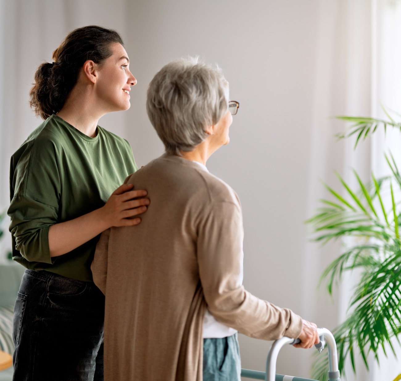 A caregiver steadying an older woman as she walks with a wheeled walker at home